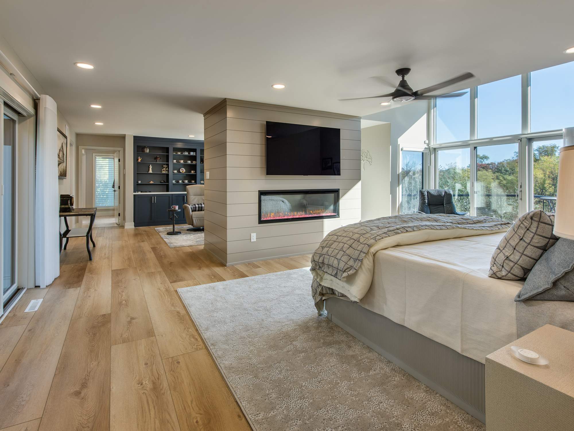 Primary bedroom with a two-sided fireplace, mounted TV, wide-plank flooring, and expansive windows