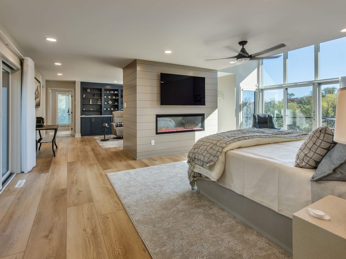Primary bedroom with a two-sided fireplace, mounted TV, wide-plank flooring, and expansive windows