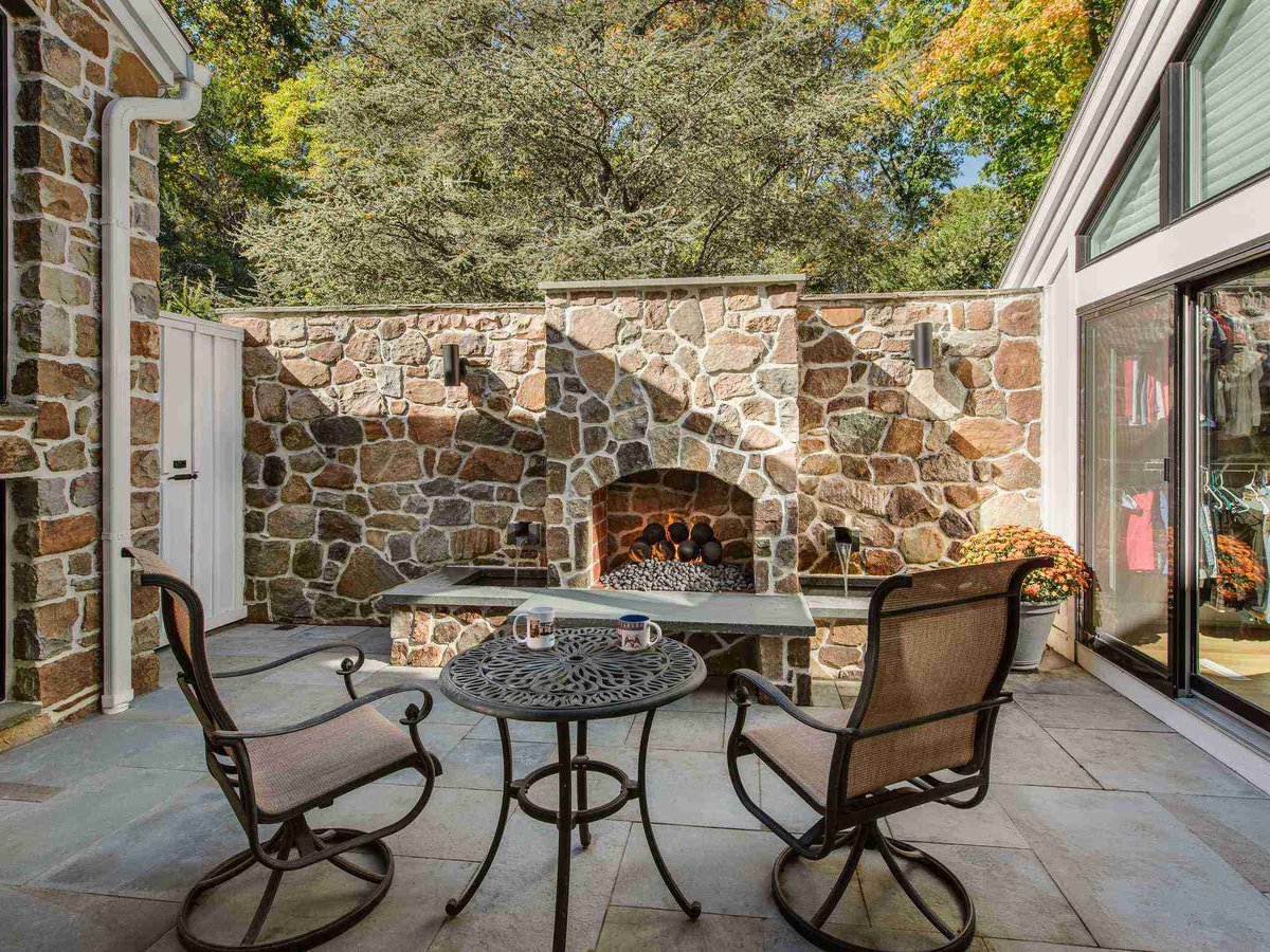 Private stone courtyard with a custom fireplace, bistro table, and swivel chairs surrounded by mature trees